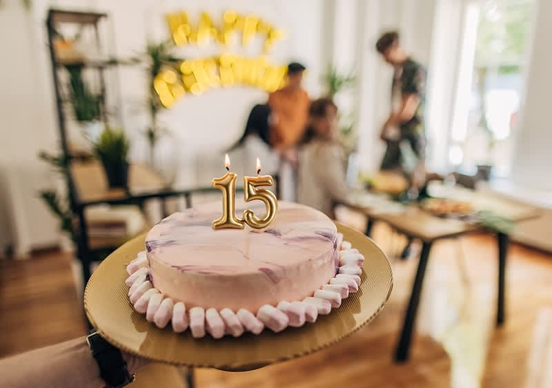 Eine Torte mit der Zahl 15 aus Kerzen darauf, umgeben von rosa und weißen Marshmallows, wird auf einem goldenen Teller gehalten. Im Hintergrund sind unscharf drei Personen und goldene Luftballons mit der Aufschrift „Party“ erkennbar