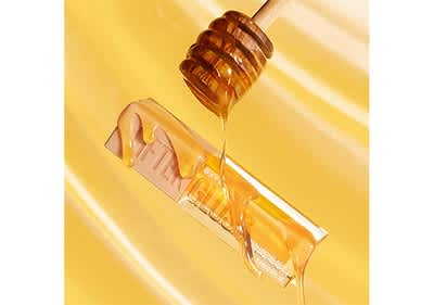 Ein goldener Honiglöffel tropft flüssigen Honig auf einen transparenten Lippenpflegestift mit der Aufschrift FTELOUS, der auf einem Hintergrund in verschiedenen Gelbtönen liegt, die an fließenden Honig erinnern