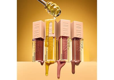 Vier Lipgloss-Flaschen mit beige Deckeln und der Aufschrift Lifter Gloss stehen nebeneinander vor einem goldenen Hintergrund, wobei ein goldener Honiglöffel von oben herabtropft und die Flaschen mit glänzendem Honig überzieht, die Flaschen sind in verschiedenen Farben: dunkelbraun, hellbraun, rosa und transparent