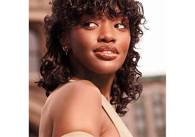 Eine Person mit lockigem, dunklem Haar trägt ein cremefarbenes Oberteil mit einem asymmetrischen Ausschnitt, im Hintergrund sind verschwommene Gebäude zu erkennen.