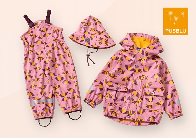 Eine Kinderregenbekleidungskollektion bestehend aus einem rosa Regenanzug, einer Regenjacke und einem Regenhut, alle mit einem bunten Schirmmuster in Gelb, Grau und Schwarz, die Jacke hat eine gelbe Innenseite und zwei Taschen, der Regenanzug hat verstellbare Träger und elastische Bündchen, der Hut hat eine Kordel zum Festziehen, oben rechts befindet sich ein orangefarbenes Quadrat mit dem weißen Schriftzug PUSBLU und zwei stilisierten Pusteblumen.