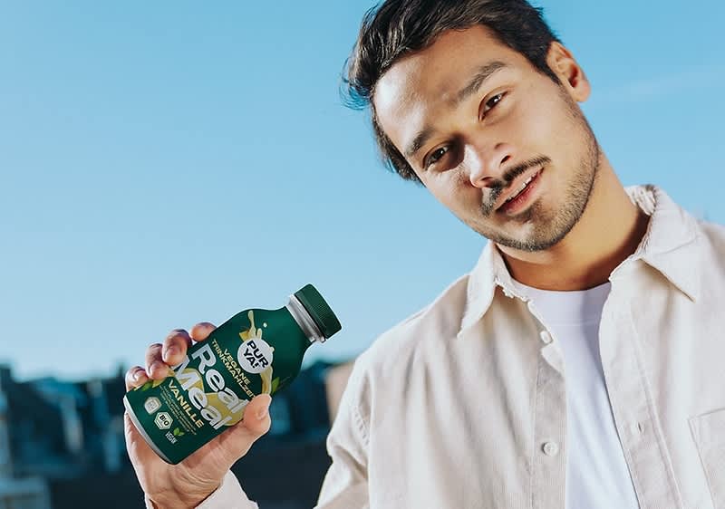 Ein Mann mit hellem Hemd hält eine grüne Flasche mit der Aufschrift Real Meal und Vanilla in der Hand, die Flasche hat einen grauen Deckel und im Hintergrund ist ein blauer Himmel zu sehen