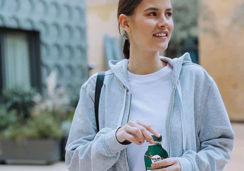 Eine Person in einem grauen Hoodie hält eine grüne Flasche mit der Aufschrift „Pure“