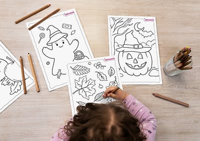 Ein Kind mit lockigem Haar und einem lila gepunkteten Pullover malt an einem Tisch mit Buntstiften auf einem Blatt Papier, das Herbstblätter zeigt, daneben liegen zwei weitere Ausmalbilder mit einem fröhlichen Geist mit Kürbis und einem Kürbis mit Hexenhut.