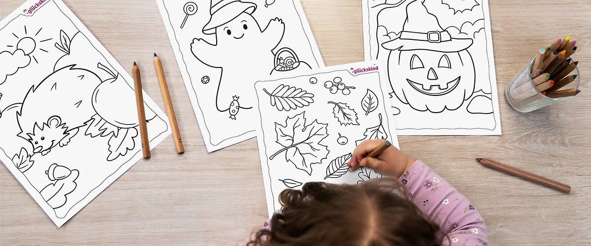 Ein Kind mit lockigem Haar und einem lila gepunkteten Pullover malt an einem Tisch mit Buntstiften auf einem Blatt Papier, das Herbstblätter zeigt, daneben liegen zwei weitere Ausmalbilder mit einem fröhlichen Geist mit Kürbis und einem Kürbis mit Hexenhut.