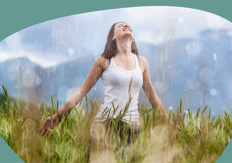 Eine Frau mit schulterlangem braunem Haar steht in einem grünen Feld mit hohem Gras, trägt ein weißes ärmelloses Oberteil und hält ihre Arme seitlich ausgestreckt, während sie den Kopf nach oben richtet, um den Regen zu genießen, im Hintergrund sind verschwommene Berge zu sehen und der Himmel ist bewölkt.