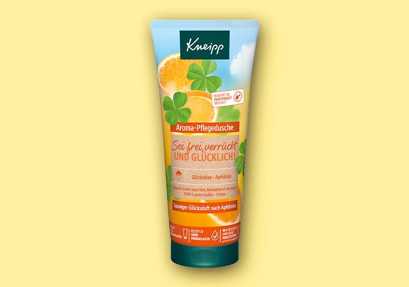 Eine orangefarbene Tube der Kneipp Aroma-Pflegedusche mit der Aufschrift „Sei frei, verrückt und glücklich“, verziert mit einem Kleeblatt und einer Orangenscheibe, darunter „Glücklicher Apfel“ und „Fruchtiger Glücksduft nach Apfel“ auf einem gelben Hintergrund