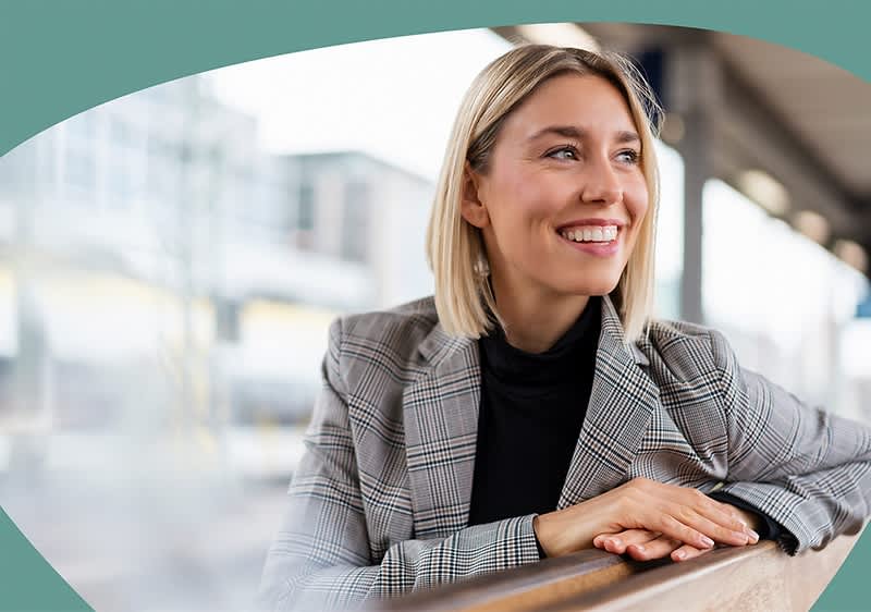 Eine Frau mit schulterlangem blondem Haar trägt einen grauen karierten Blazer über einem schwarzen Rollkragenpullover und lehnt sich mit verschränkten Armen auf eine Holzbank in einem modernen, lichtdurchfluteten Raum mit großen Fenstern