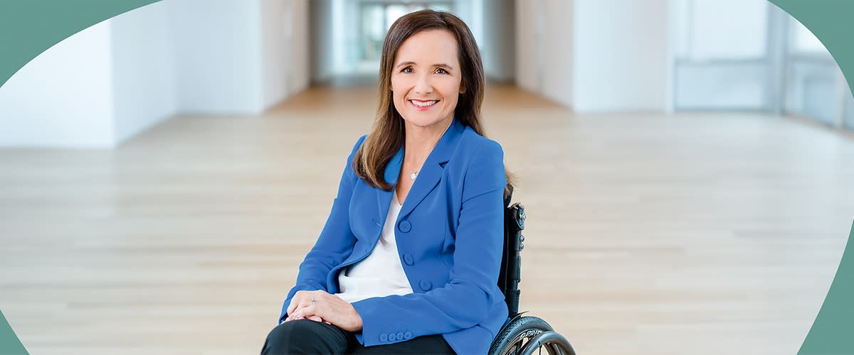 Mirjam Kottmann mit langen braunen Haaren sitzt in einem Rollstuhl, sie trägt einen blauen Blazer über einem weißen Oberteil und eine schwarze Hose, der Hintergrund zeigt einen hellen Raum mit Holzboden und weißen Wänden.