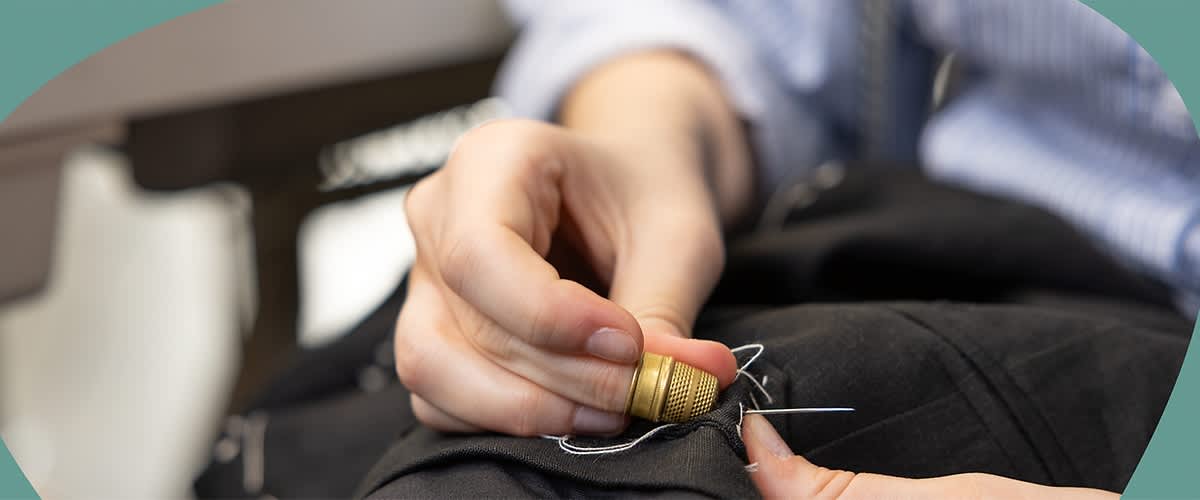 Eine Nahaufnahme einer Hand, die einen Fingerhut trägt und mit einer Nadel und weißem Faden schwarzen Stoff näht, im Hintergrund ist unscharf ein weiteres Stück Stoff zu erkennen.