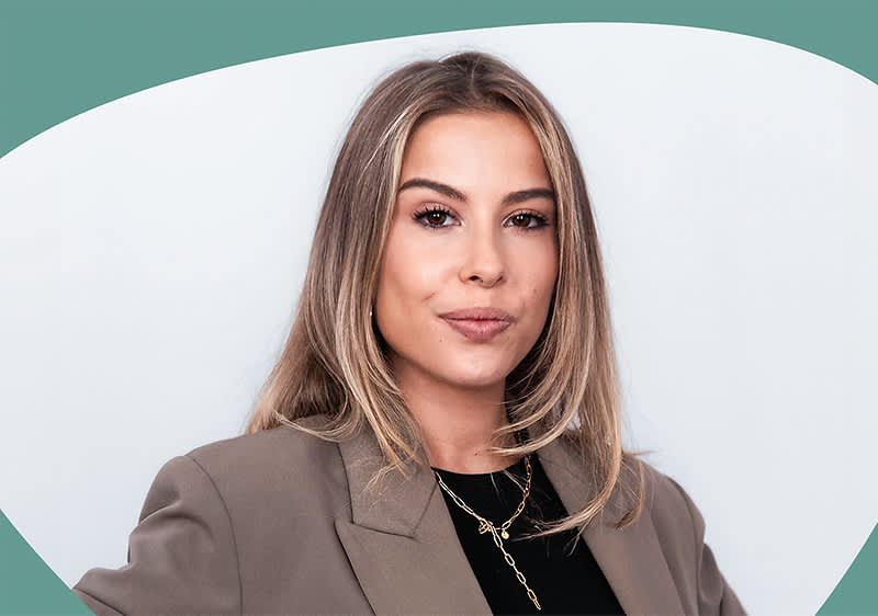 Miriam Weilmünster trägt einen grauen Blazer und ein schwarzes Oberteil, dazu eine goldene Halskette mit einem Anhänger, der aus mehreren Kettengliedern besteht, der Hintergrund ist in einem hellen Grauton gehalten und oben ist ein geschwungener grüner Bereich sichtbar.