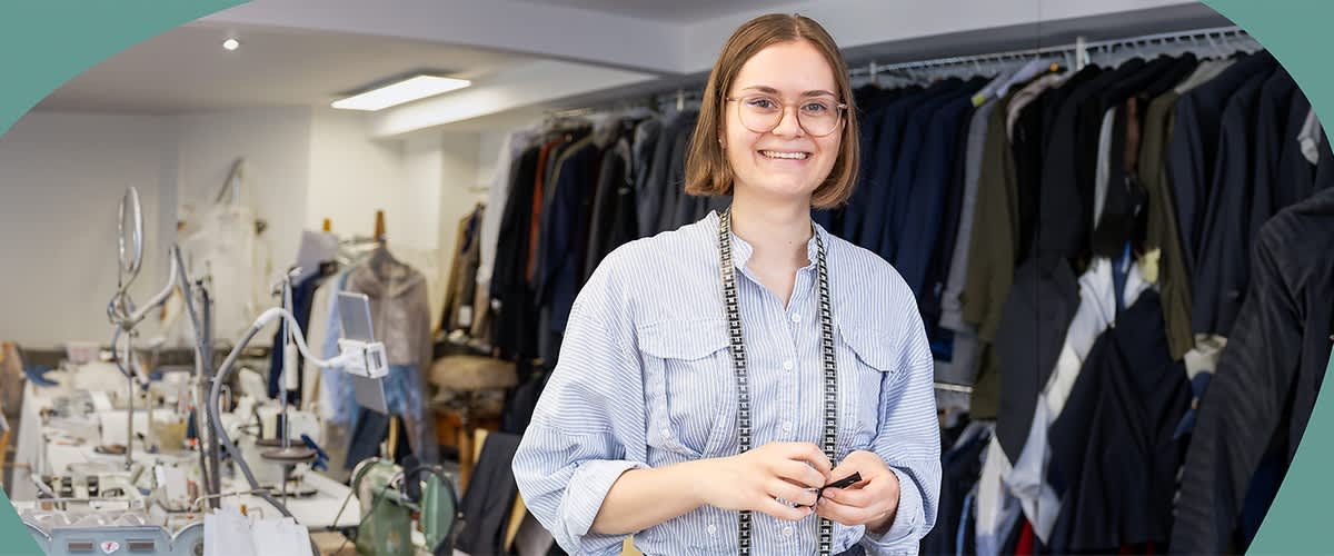 Pauline Unkelbach trägt ein hellblaues Hemd und eine schwarze Hose, um den Hals hängt ein Maßband, im Hintergrund sind zahlreiche Kleidungsstücke auf Kleiderbügeln zu sehen, die an einer Kleiderstange hängen, auf einem Tisch liegen Nähutensilien wie eine Schere und ein grünes Nähmaschinenmodell.
