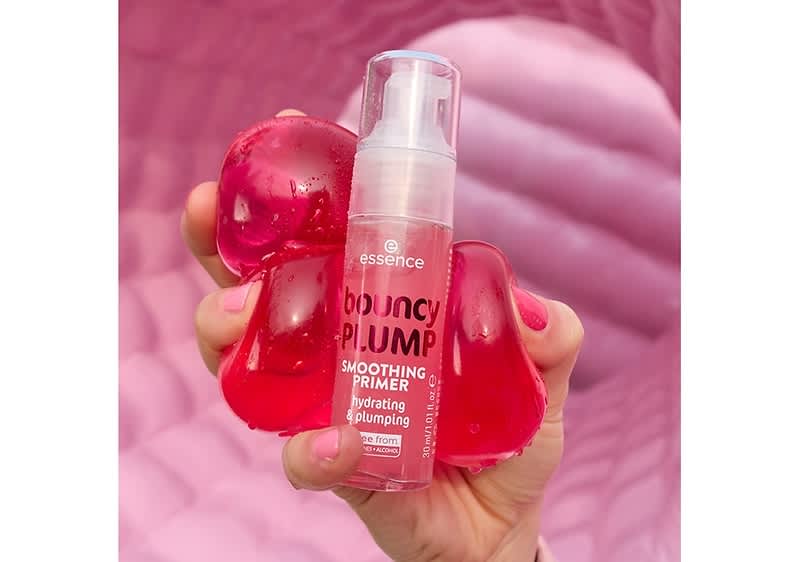 Eine Hand mit pink lackierten Fingernägeln hält eine durchsichtige Flasche mit der Aufschrift „essence bouncy PLUMP SMOOTHING PRIMER Hydrating & Plumping“ und „vegan formula“ in der Mitte, umgeben von drei pinkfarbenen, tropfenförmigen Objekten vor einem Hintergrund in verschiedenen Pinktönen.