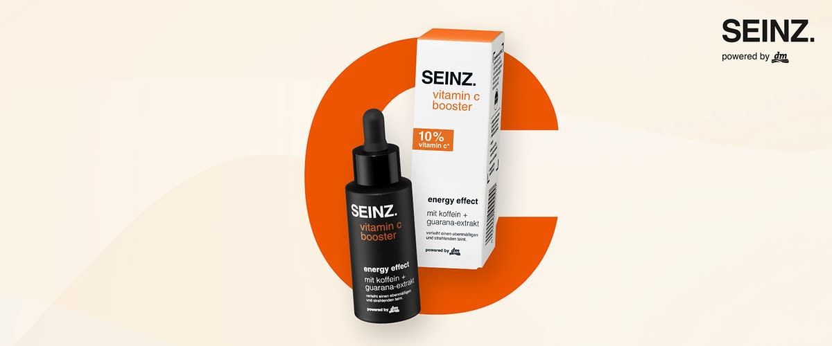 Eine Produktabbildung mit einer schwarzen Flasche und einer weißen Verpackung, beide mit der Aufschrift SEINZ. vitamin c booster, die Flasche trägt zusätzlich die Worte energy effect mit koffein + guarana-extrakt und powered by dm, die Verpackung zeigt 10% vitamin c* und energy effect mit koffein + guarana-extrakt, im Hintergrund ein großes oranges C und oben rechts der Schriftzug SEINZ. powered by dm