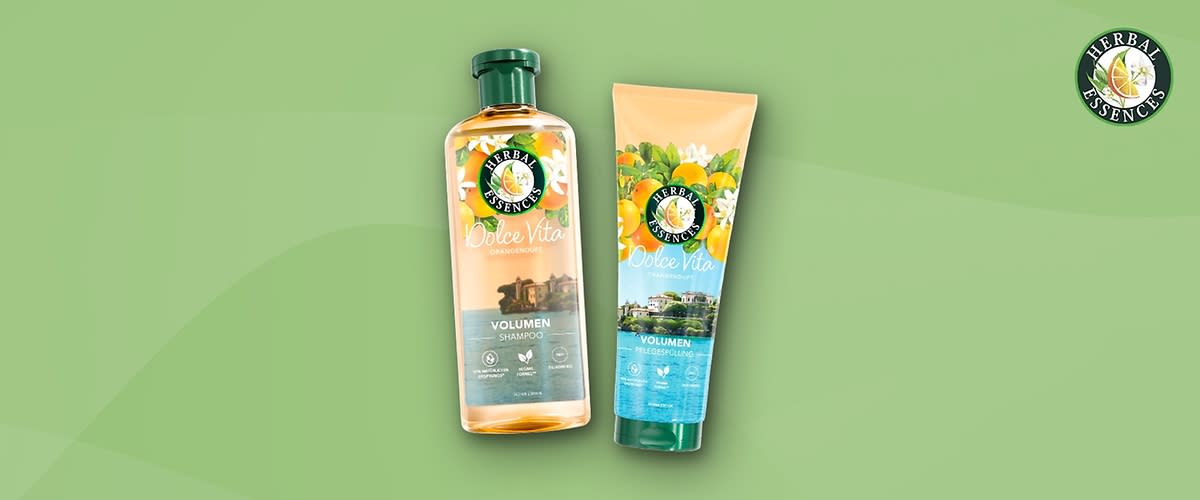 Zwei Produktverpackungen von Herbal Essences auf einem grünen Hintergrund, links eine durchsichtige Flasche mit grünem Deckel und orangefarbener Flüssigkeit, darauf das Logo mit einer Rose und der Text Herbal Essences Dolce Vita Orangenblüte Volumen Shampoo, darunter Symbole für vegan und recycelbar, rechts eine Tube mit grünem Deckel und einem Bild von Orangenblüten und Orangen, darauf ebenfalls das Logo und der Text Dolce Vita Volumen