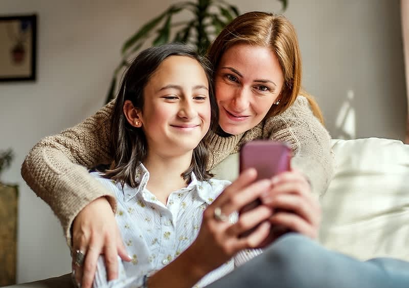 Zwei Personen sitzen auf einem Sofa, die vordere Person hält ein Smartphone in der Hand, während die hintere Person sie umarmt
