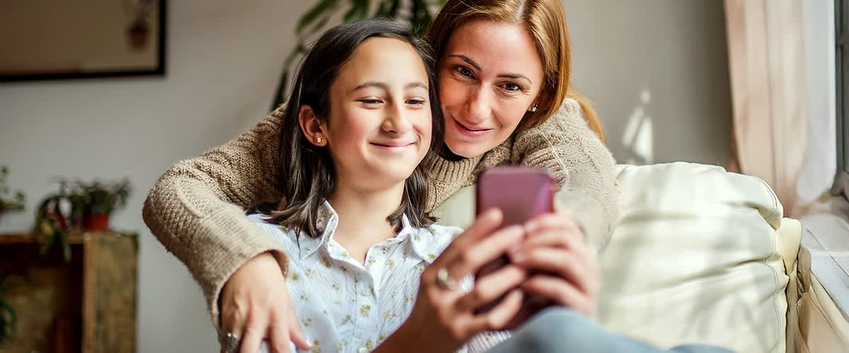 Zwei Personen sitzen auf einem Sofa, die vordere Person hält ein Smartphone in der Hand, während die hintere Person sie umarmt