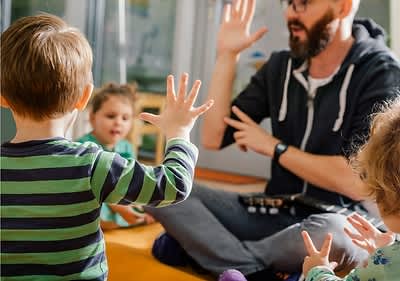 Ein Kind in einem gestreiften Pullover hebt die Hand, während ein Erwachsener mit verschwommenem Gesicht im Hintergrund ebenfalls die Hand hebt