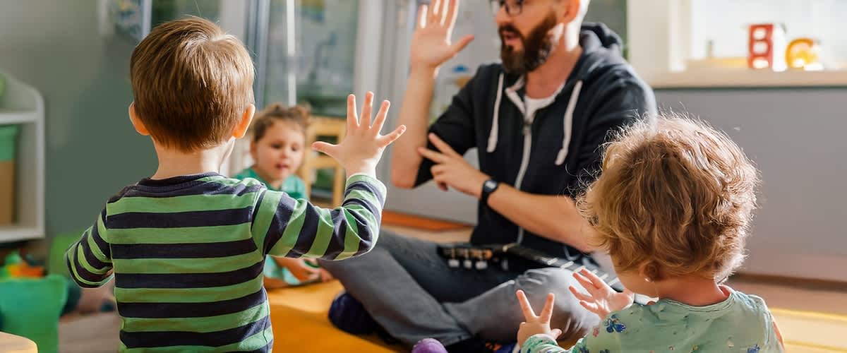 Ein Kind in einem gestreiften Pullover hebt die Hand, während ein Erwachsener mit verschwommenem Gesicht im Hintergrund ebenfalls die Hand hebt