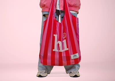Mareike in blauer Jeans und weißen Turnschuhen hält eine rot-weiß gestreifte Einkaufstasche mit der Aufschrift „hi“ und einem Smiley