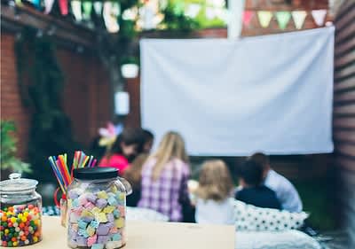 Ein Glas mit bunten Süßigkeiten und Trinkhalmen steht im Vordergrund, im Hintergrund sitzen vier Personen vor einer weißen Leinwand im Garten