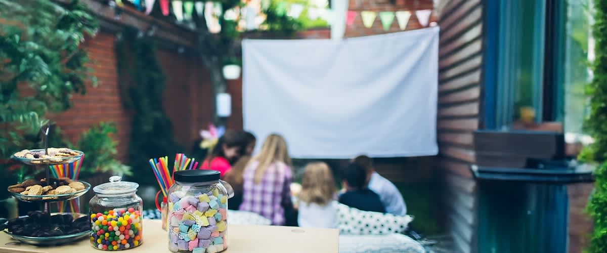 Ein Glas mit bunten Süßigkeiten und Trinkhalmen steht im Vordergrund, im Hintergrund sitzen vier Personen vor einer weißen Leinwand im Garten