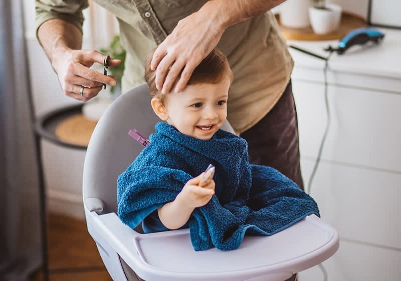 Ein Kleinkind sitzt in einem Hochstuhl und trägt einen blauen Umhang, während eine Person mit einer Schere die Haare des Kindes schneidet