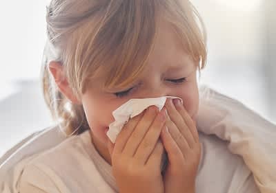 Eine Person mit blonden Haaren trägt ein weißes Oberteil und putzt die Nase mit einem Taschentuch.