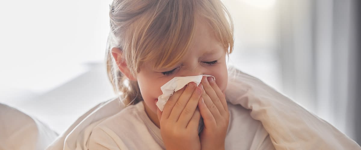 Eine Person mit blonden Haaren trägt ein weißes Oberteil und putzt die Nase mit einem Taschentuch.