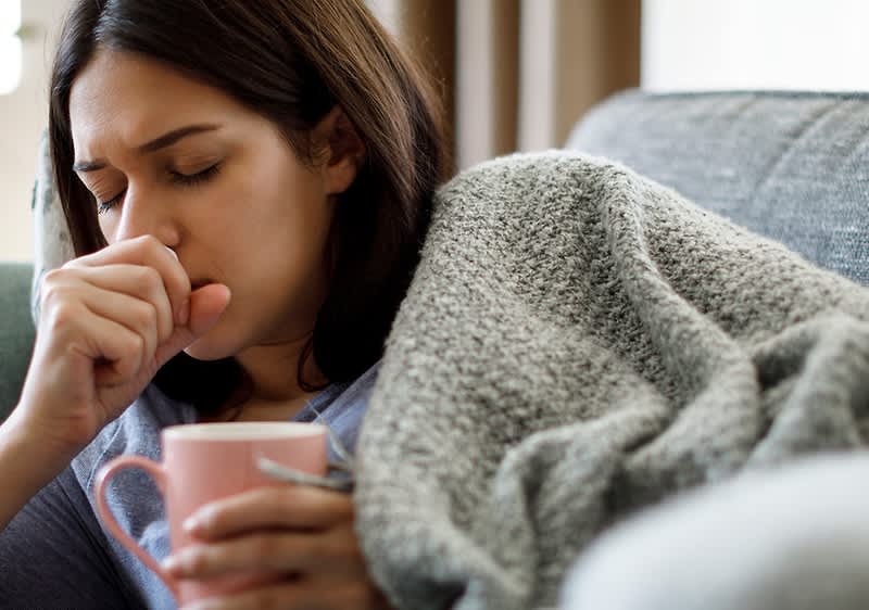 Eine hustende Person sitzt auf einem Sofa, in eine graue Decke gehüllt, und hält eine rosa Tasse in der Hand. Die andere Hand hält sie vor ihren Mund.