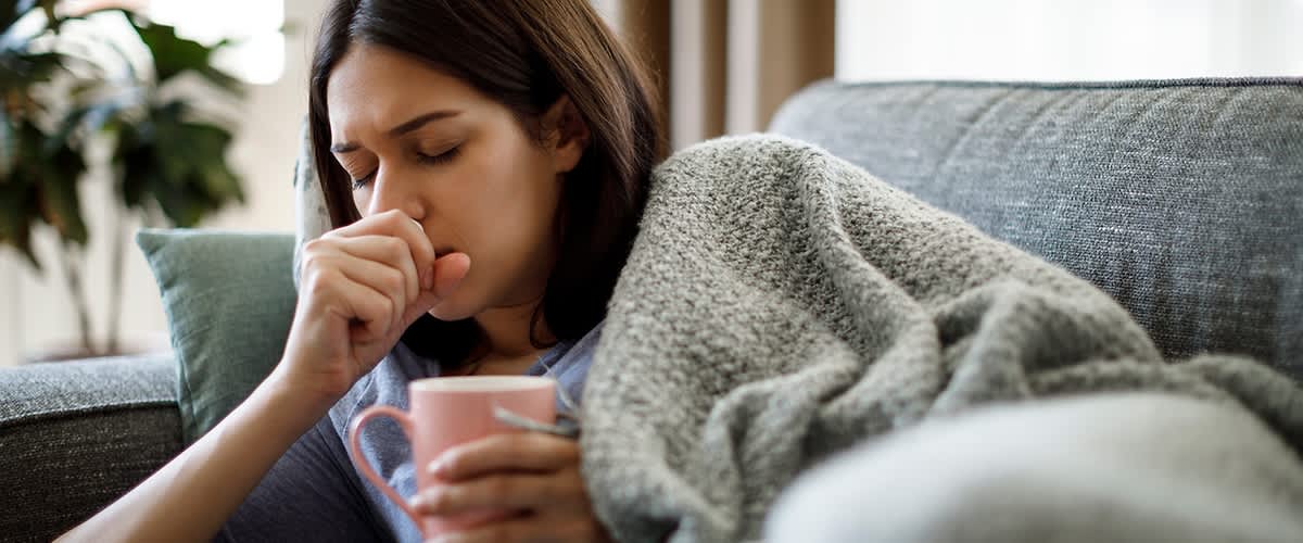 Eine hustende Person sitzt auf einem Sofa, in eine graue Decke gehüllt, und hält eine rosa Tasse in der Hand. Die andere Hand hält sie vor ihren Mund.