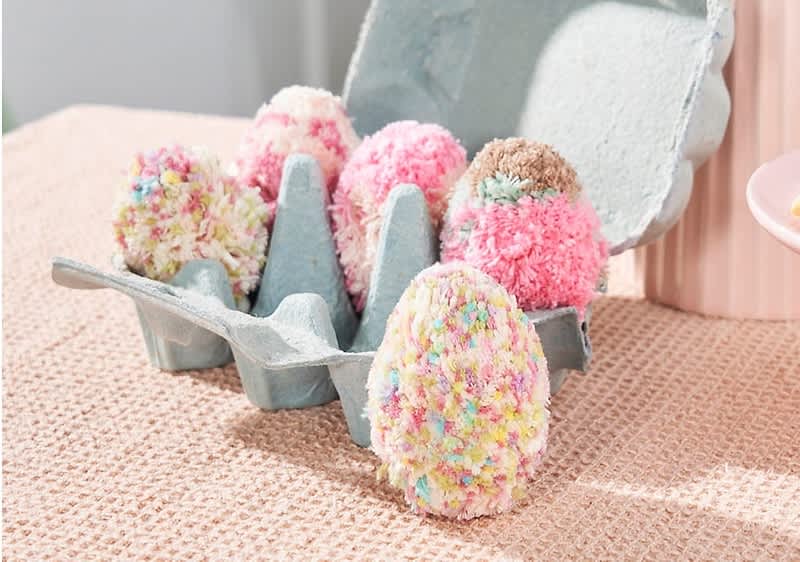 Vier bunte, flauschige Eier in einer grauen Eierkartonverpackung auf einem beigen Tisch