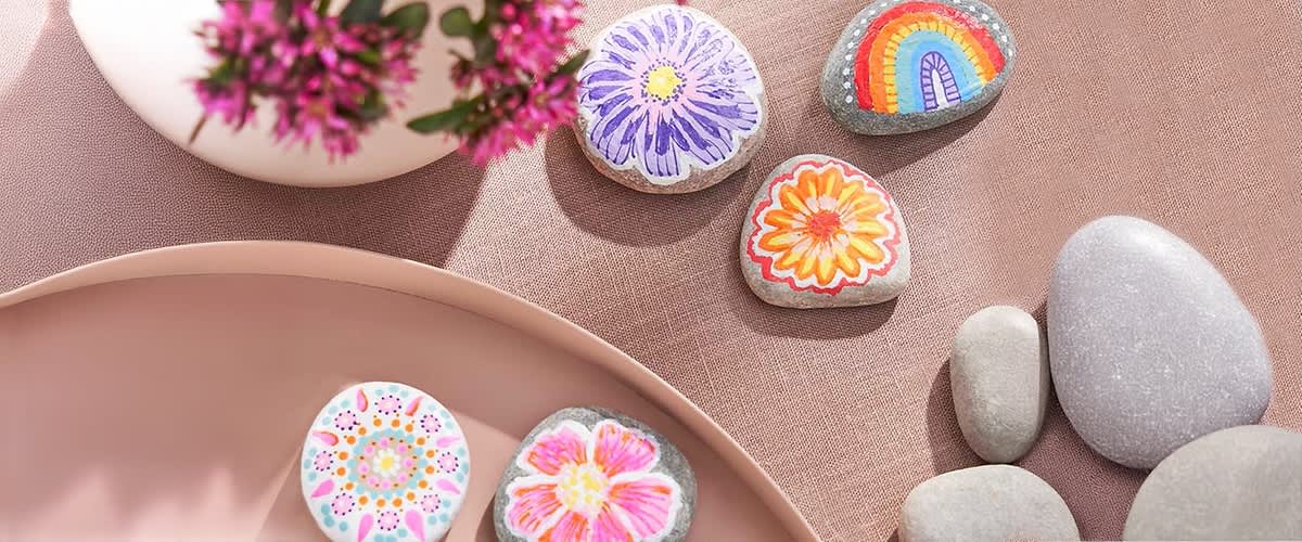 Fünf bemalte Steine mit bunten Mustern liegen auf einem rosa Stoff, darunter ein Stein mit lila Blume, ein Stein mit Regenbogen, ein Stein mit orangefarbener Blume, ein Stein mit Mandala und ein Stein mit rosa Blume