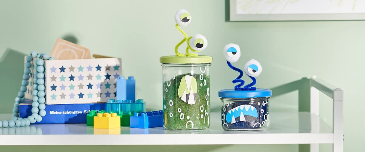 Zwei Gläser mit bunten Deckeln, dekoriert mit Augen und Monstergesichtern, stehen auf einem Regal neben bunten Lego-Steinen