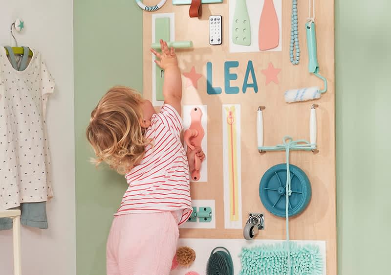 Ein kleines Kind mit lockigem Haar und gestreiftem T-Shirt spielt an einer grünen Wand mit verschiedenen Gegenständen, darunter ein Roller, Bürsten, ein Lichtschalter und der Name „LEA“ in bunten Buchstaben