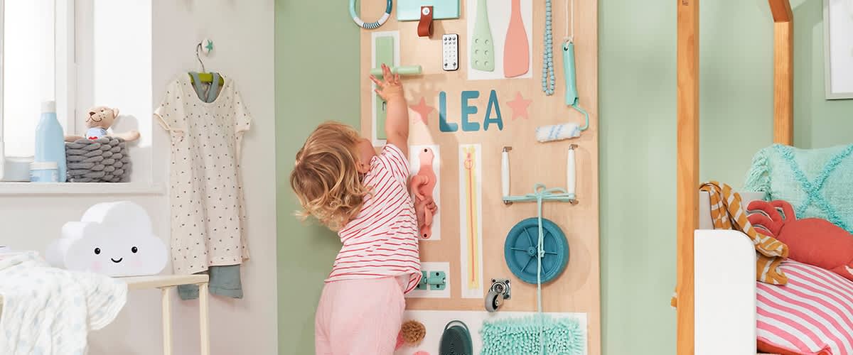 Ein kleines Kind mit lockigem Haar und gestreiftem T-Shirt spielt an einer grünen Wand mit verschiedenen Gegenständen, darunter ein Roller, Bürsten, ein Lichtschalter und der Name „LEA“ in bunten Buchstaben
