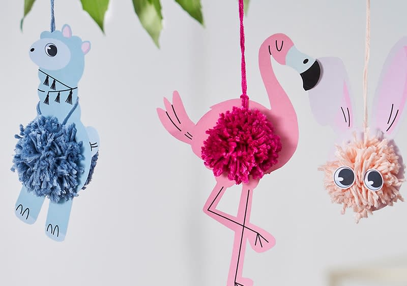 Drei Papierfiguren hängen an Fäden, ein blaues Lama mit einem blauen Pompon, ein rosa Flamingo mit einem rosa Pompon und ein rosa Hase mit einem cremefarbenen Pompon