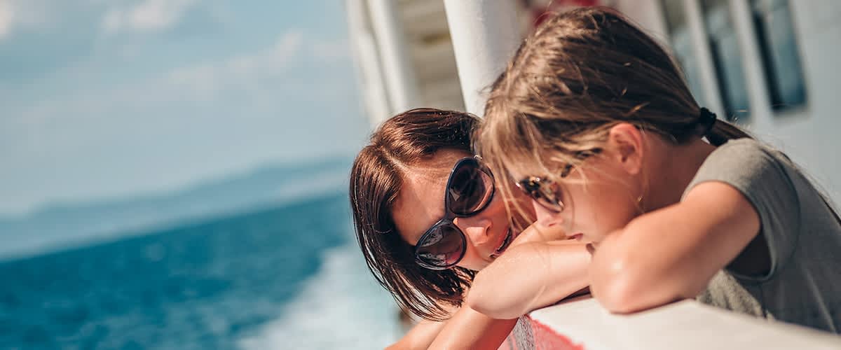 Eine Frau mit braunen Haaren und Sonnenbrille lehnt sich lächelnd über die Reling eines Schiffes, während im Hintergrund das Meer zu sehen ist
