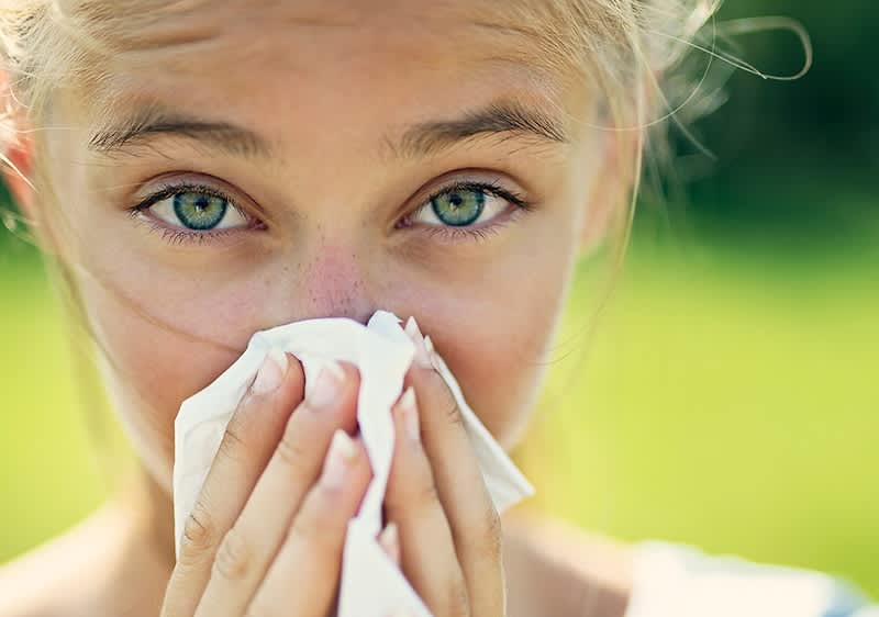 Eine Person mit hellblondem Haar putzt sich die Nase mit einem Taschentuch vor einem grünen Hintergrund
