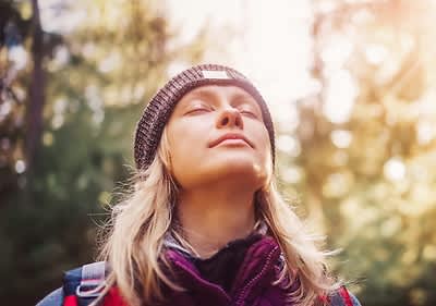 Eine Person mit blonden Haaren trägt eine Jacke und eine braune Strickmütze, im Hintergrund sind unscharfe Bäume und Sonnenlicht zu sehen
