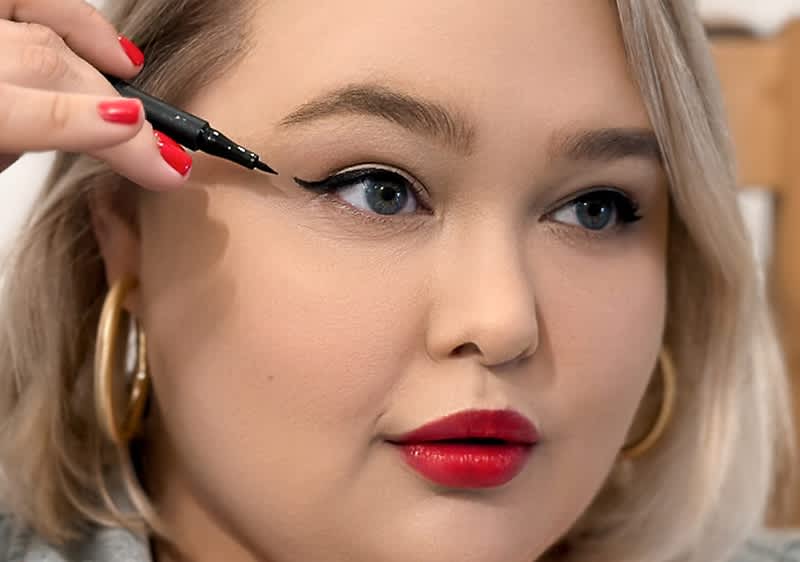 Lidstrich auftragen: Eine Person mit blonden Haaren trägt große goldene Ohrringe und hält eine schwarze Haarspange in der Hand