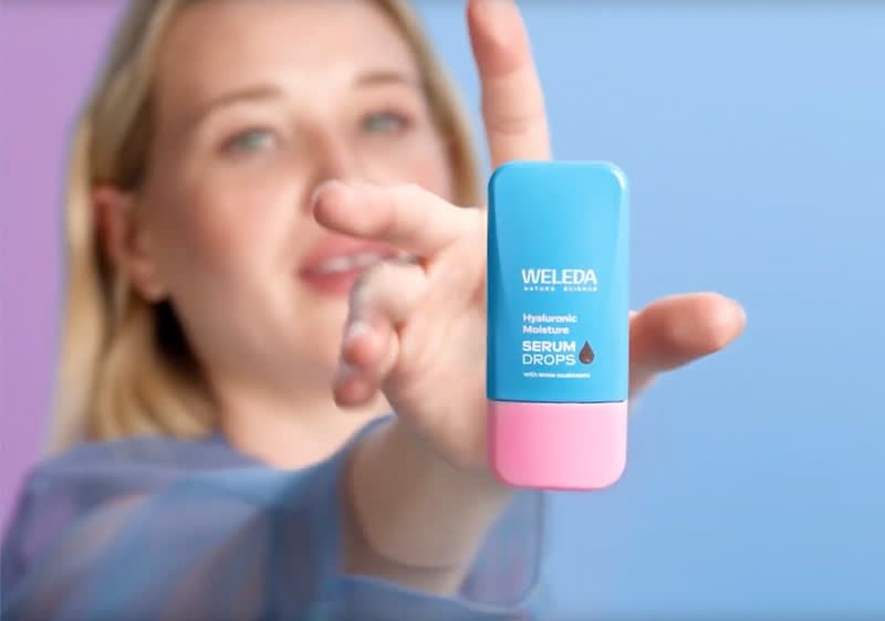 Eine Hand hält eine blaue Flasche mit pinkem Verschluss, darauf steht „Weleda Hydrating Serum Drops“