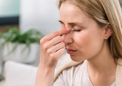 Eine Frau mit blonden Haaren hält ihre Hand an die Nase, im Hintergrund ist eine grüne Pflanze in einem weißen Topf erkennbar