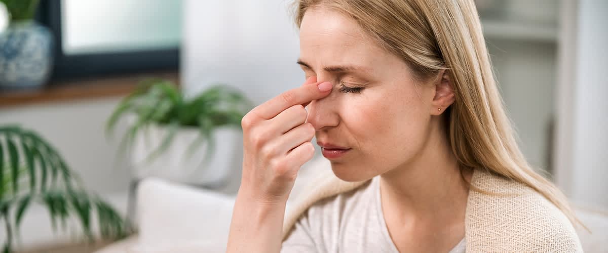Eine Frau mit blonden Haaren hält ihre Hand an die Nase, im Hintergrund ist eine grüne Pflanze in einem weißen Topf erkennbar