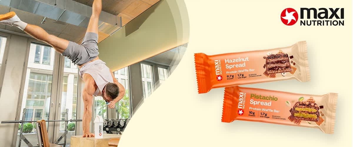 Ein Mann in sportlicher Kleidung macht einen Handstand auf einem Holzkasten, daneben zwei Protein-Waffelriegel von Maxi Nutrition, einer mit Haselnusscreme und einer mit Pistaziencreme, beide mit Nährwertangaben auf der Verpackung, oben rechts das Logo von Maxi Nutrition