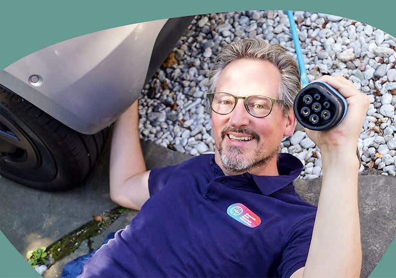 Dr. Eckart von Hirschhausen im dunkelblauen Poloshirt liegt auf dem Boden neben einem Auto und hält einen Ladestecker für Elektrofahrzeuge in der Hand