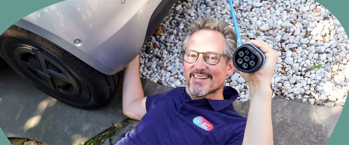 Dr. Eckart von Hirschhausen im dunkelblauen Poloshirt liegt auf dem Boden neben einem Auto und hält einen Ladestecker für Elektrofahrzeuge in der Hand