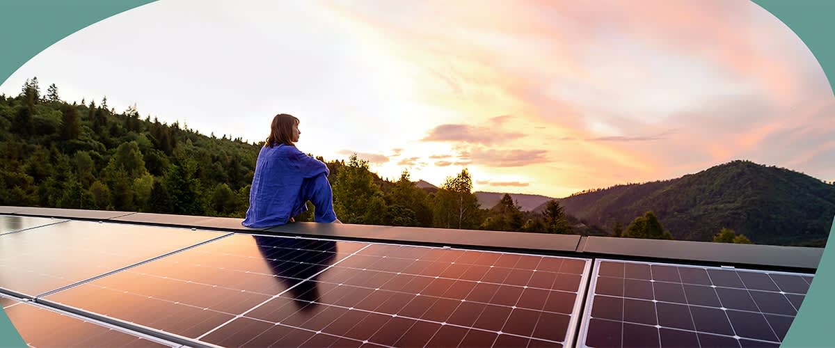Eine Person in blauer Kleidung sitzt auf einem Solarpanel und blickt in die Ferne, umgeben von einer bewaldeten Landschaft bei Sonnenuntergang