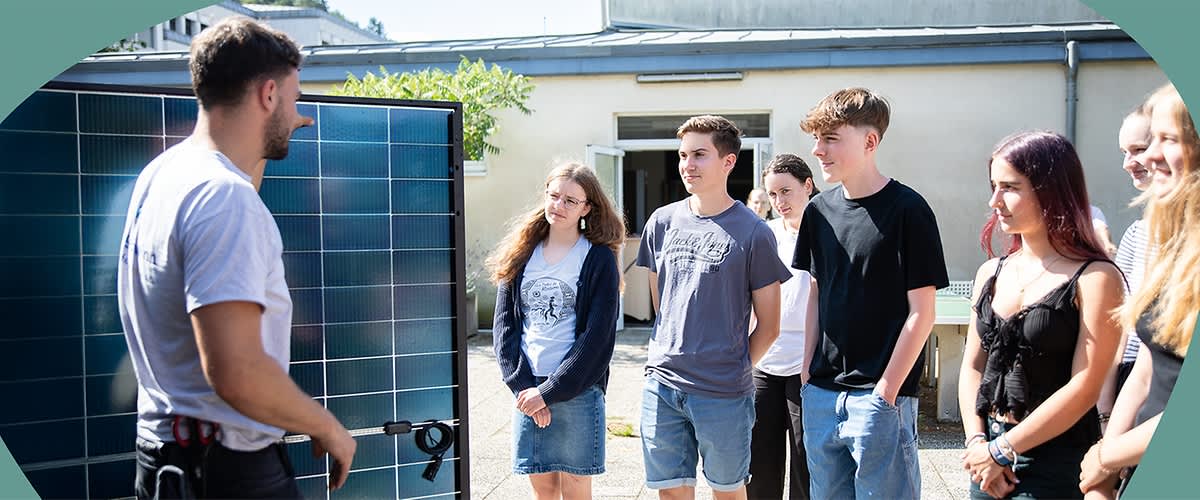 Eine Gruppe von fünf jungen Erwachsenen steht im Freien und hört einem Mann zu, der ihnen ein Solarpanel erklärt