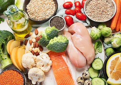 Verschiedene gesunde Lebensmittel wie Hähnchenbrust, Lachs, Brokkoli, Blumenkohl, Tomaten, Gurken, Karotten, Rosenkohl, Quinoa, Haferflocken, Chiasamen, Mandeln, Linsen, Wachteleier, Birne, Zitrone und eine Flasche Olivenöl